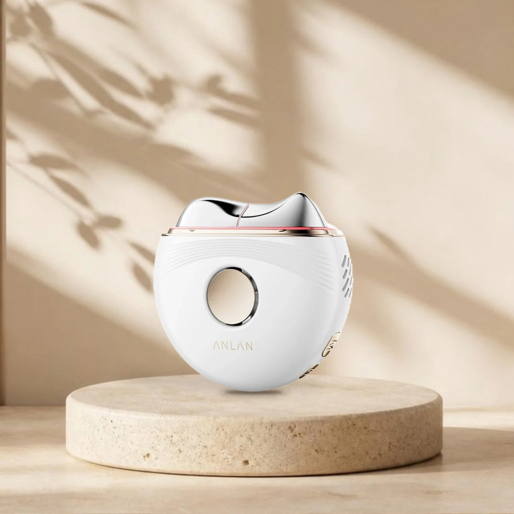 Gua sha électrique chaud et froid ANLAN sur une plinthe en travertin, lumière naturelle et ombres de feuillage