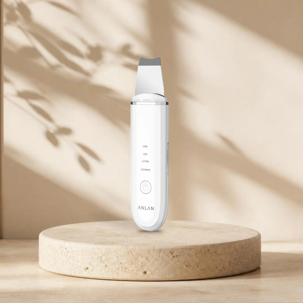 Spatule ultrasonique visage ANLAN blanche sur une plinthe en travertin, lumière naturelle et ombres de feuillage