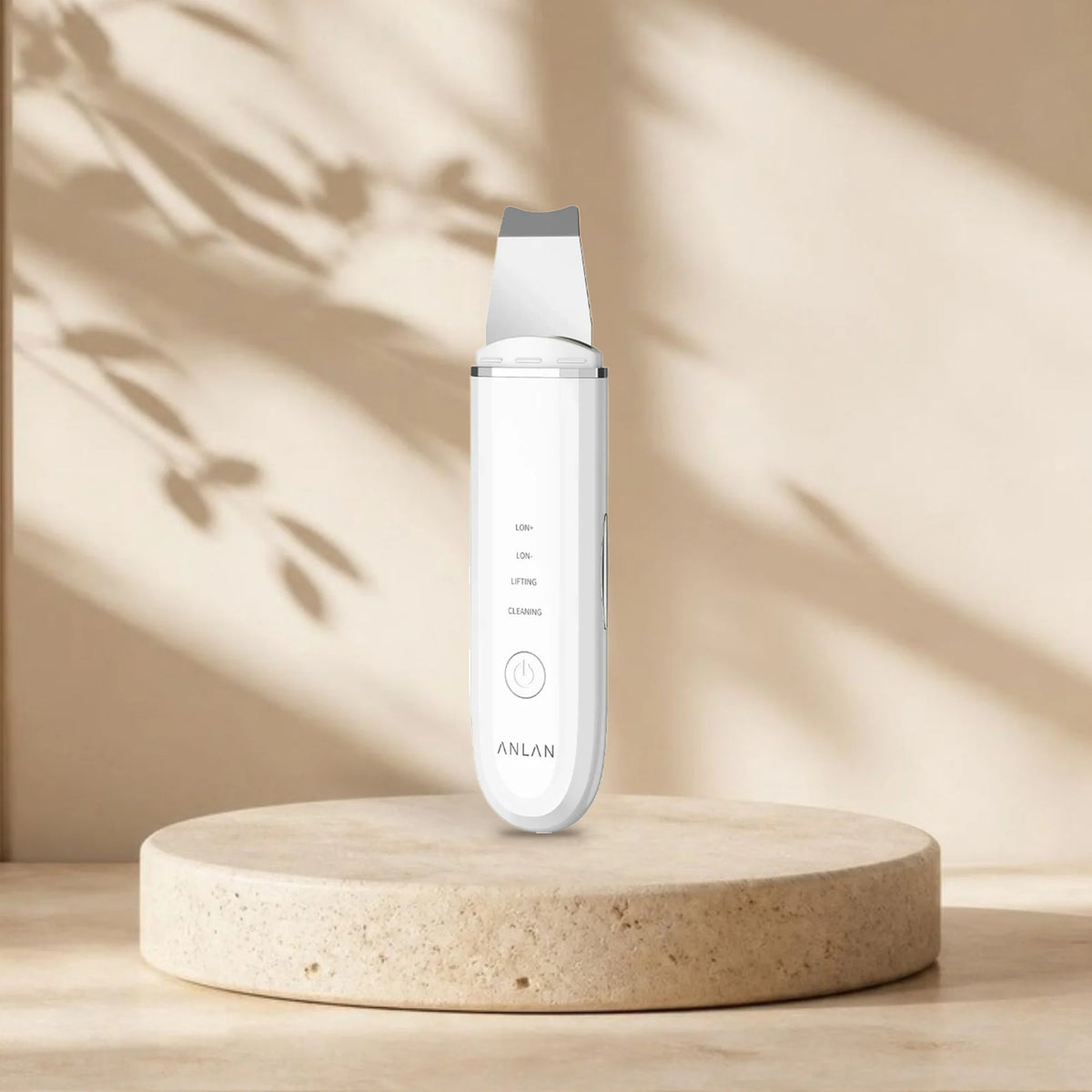 Spatule ultrasonique visage ANLAN blanche sur une plinthe en travertin, lumière naturelle et ombres de feuillage